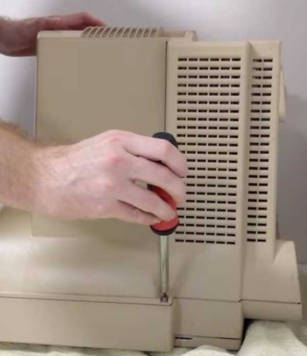 A Macintosh LC575 with the screen facing down and a hand holding a screwdriver over one of the screws attaching the case to the front panel